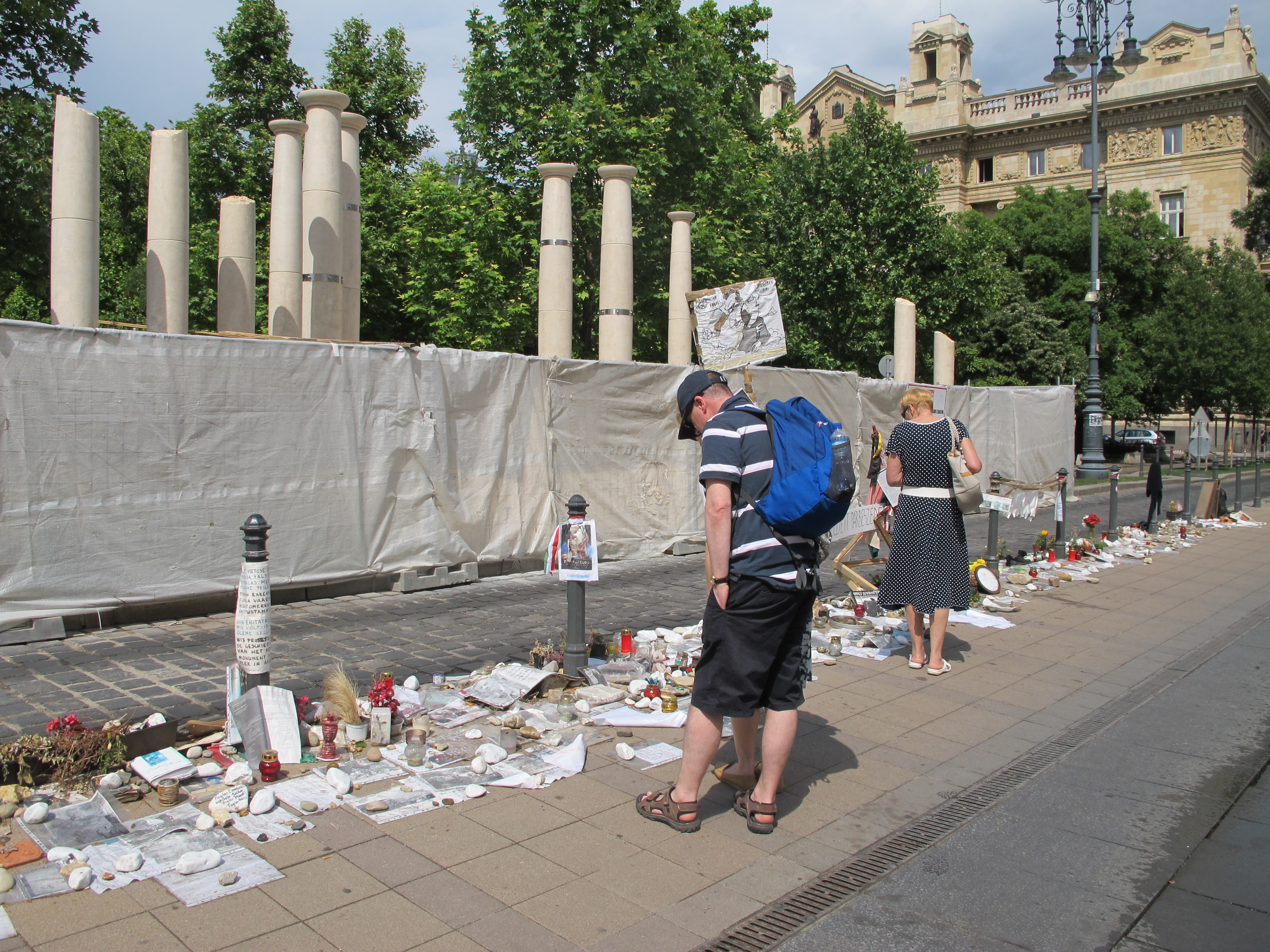 Passersby look at the Holocaust-related and other memorabilia left by citizens protesting the monument to the 1944 German occupation, under construction behind sheeting across the street in downtown Budapest. (Ruth Ellen Gruber)