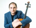 Wearing a blue jacket, Julian Schwarz stands with his arm around a cello.