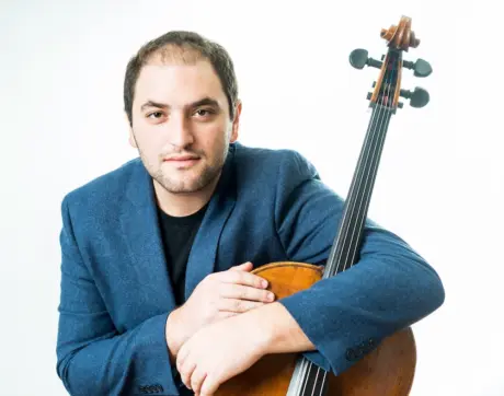 Wearing a blue jacket, Julian Schwarz stands with his arm around a cello.