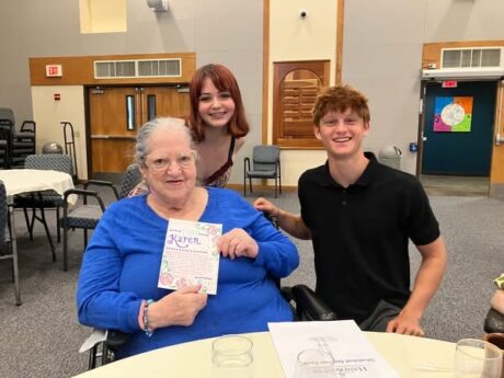 An older woman seated holds a card. Behind and to her left are two teenagers.
