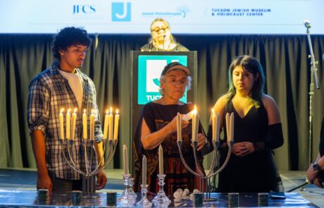 An older woman lights a candle on a candelabra. To her left and right are a teenage girl and boy. A woman stands at a podium behind the candlelighting scene.