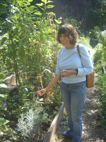 A woman dressed in jeans and a light blue top stands next to green plants in a garden plot.