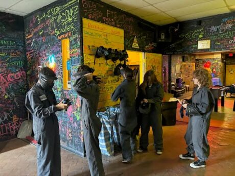 Teens dressed in coveralls put on facemasks inside a room with black walls covered in colorful graffiti.