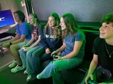 Bathed in green light from a video screen, five teens sit on bench, some holding gaming consoles.
