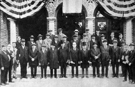 Men in suits and hats stand in front of a new synagogue building hung with bunting, in 1924.