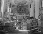 Interior of a church piled high with crates and bundles of looted items, dwarfing a soldier standing in their midst.