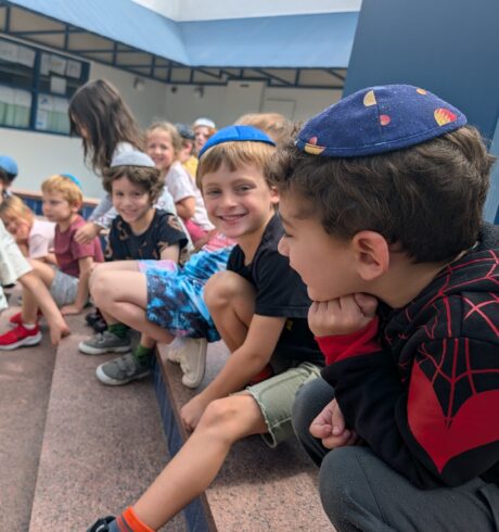 Smiling kindergarten boys wearing kippahs and girls sit on steps.